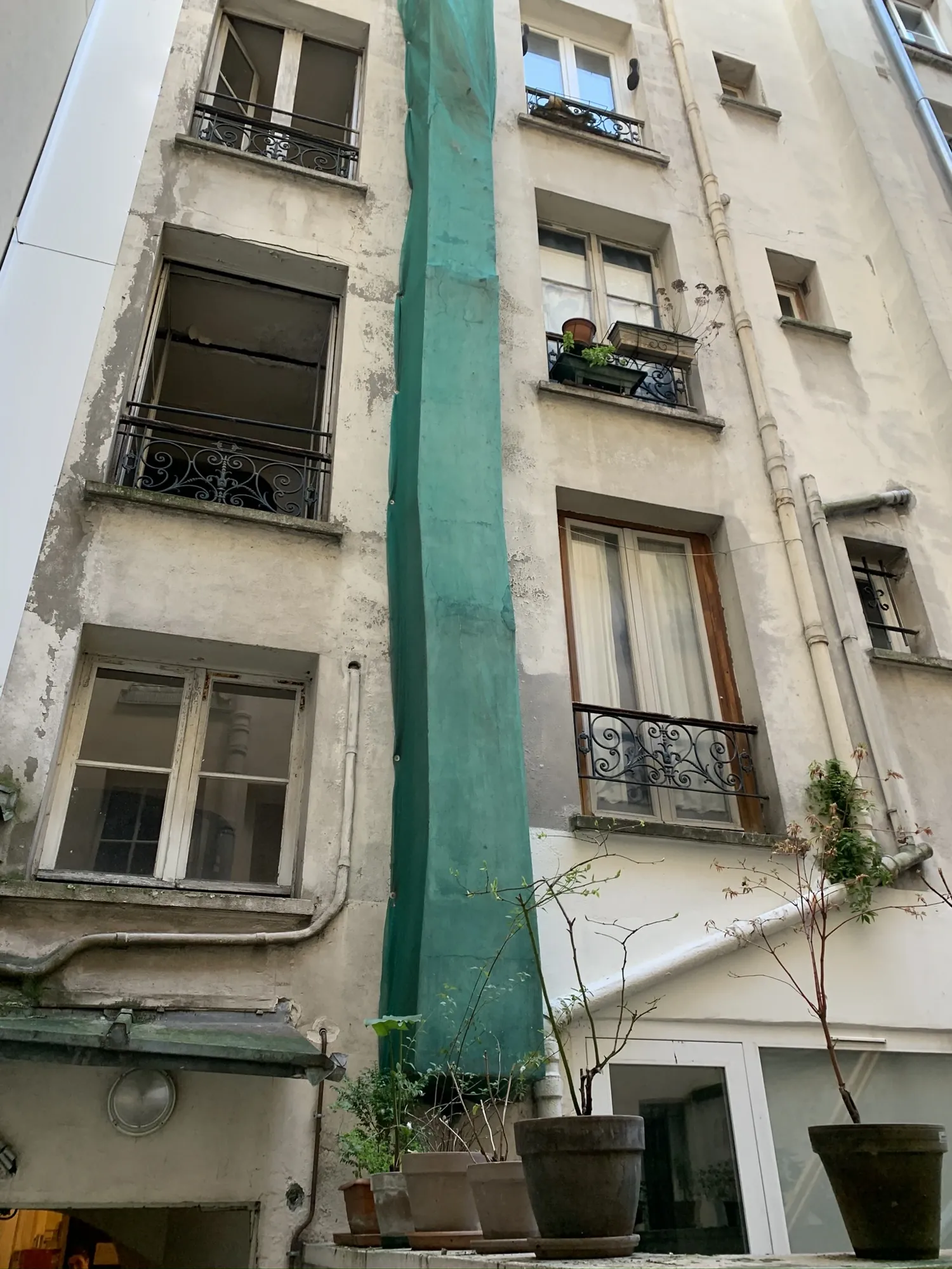 Façade sur cour d'un immeuble de 1920 à Paris 14ème avant ravalement : enduit friable et conduit de cheminée dégradé protégé par un filet vert Façade sur cour d'un immeuble de 1920 à Paris 14ème avant ravalement : enduit friable et conduit de cheminée dégradé protégé par un filet vert